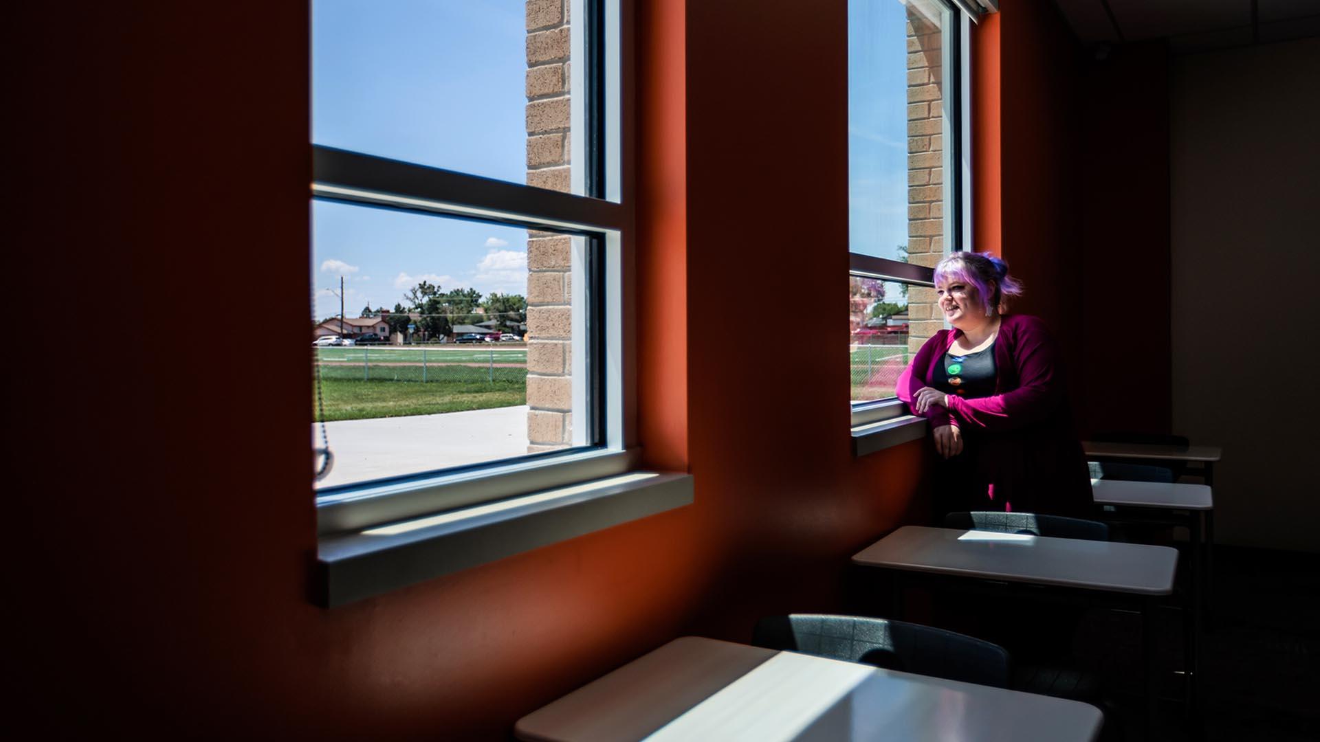 Photo of MSU Denver alumna Hayley Conley, who was recently promoted to chair of the Mathematics Department at Alameda International Jr./Sr. High in Lakewood after two years of teaching, in a dark classroom standing next to a window with bright light from outdoors. 