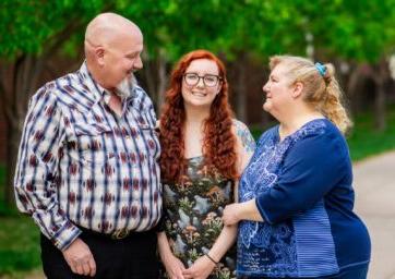 A female graduate standing with her parents outside on campus at First Generation Graduation